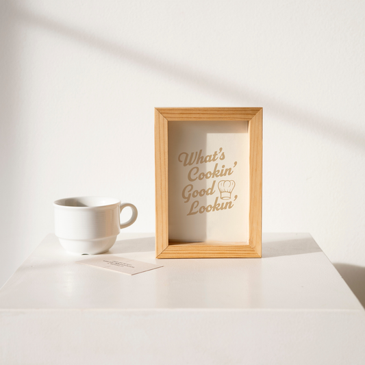 Small "What's Cookin' Good Lookin'" Kitchen Wall Art Print in tan on warm off-white. Framed cute wall decor on a clean white table.