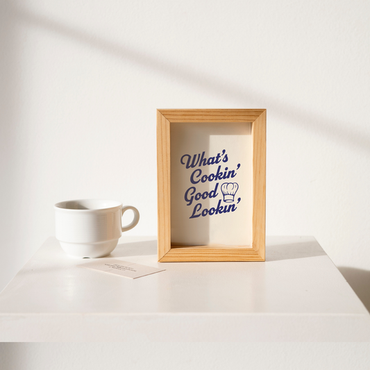 Small "What's Cookin' Good Lookin'" Kitchen Wall Art Print in blue on warm off-white. Framed cute wall decor on a clean white table.